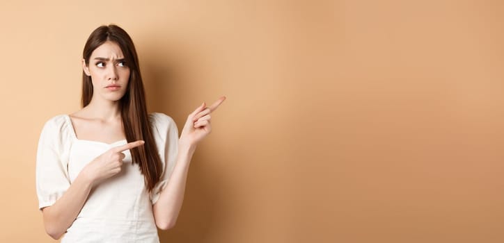 Hesitant young woman look unsure, pointing fingers right while making decision, frowning with puzzled face, standing on beige background.