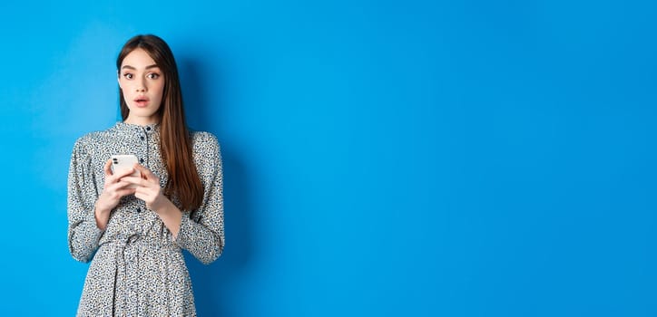 Woman look surprised after using mobile phone, standing on blue background.