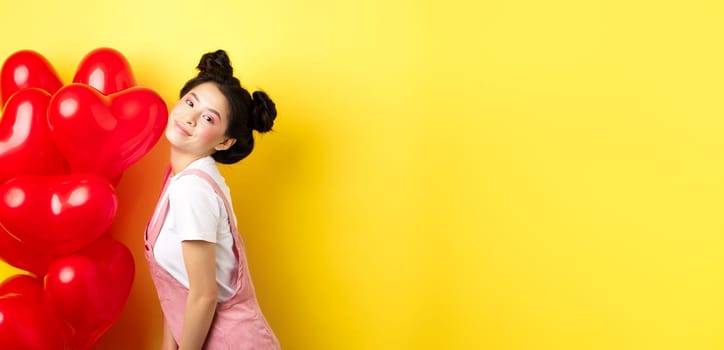 Valentines day concept. Stylish teenage asian girl posing near red hearts balloons, wear outfit for romantic date, standing happy on yellow background.