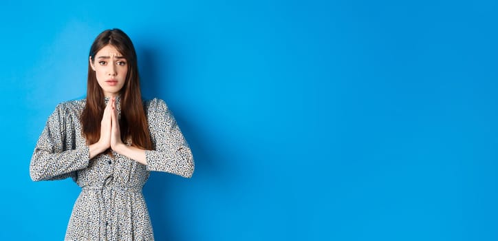 Sad girl asking for help, begging you with troubled face expression, standing in dress on blue background.