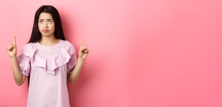 Sad pitiful asian girl sulking, pointing and looking up with disappointed face, feeling upset, standing against pink background.