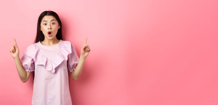 Excited asian teen girl pointing fingers up, saying wow and showing promo deal, standing in dress against pink background.