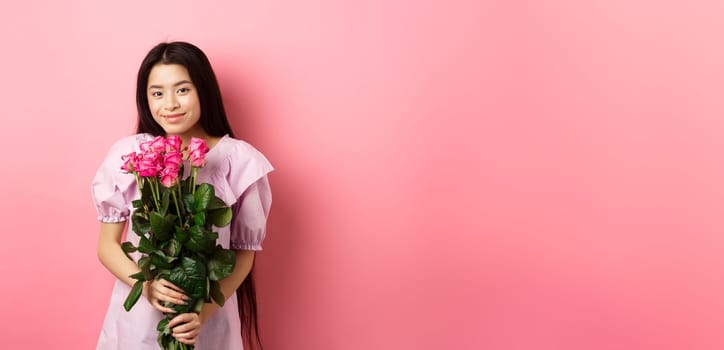 Romantic tender asian girl holding bouquet of roses, smiling cute at camera, having valentines date with lover, wearing dress, pink background.