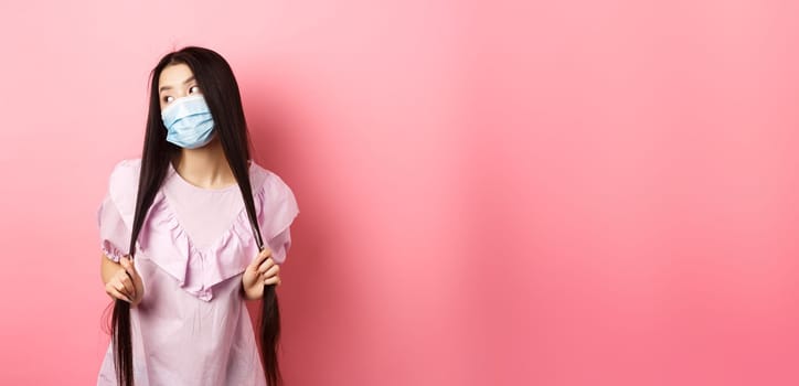 Healthy people and covid-19 pandemic concept. Stylish japanese girl in medical mask posing, looking aside and playing with long hair, standing against pink background.
