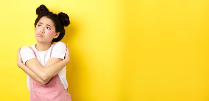 Sad lonely asian girl looking aside with upset and gloomy face, hugging herself, dreaming of love and boyfriend, yellow background.