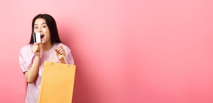 Shopping. Excited beautiful asian woman buying in stores, holding paper bag and kissing plastic credit card, standing against pink background.