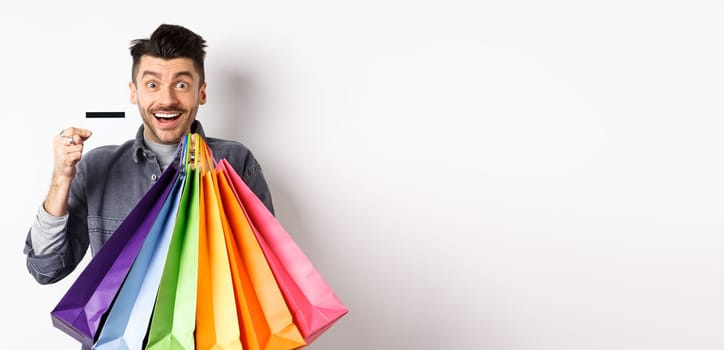 Happy guy shopaholic holding colorful shopping bags and showing plastic credit card, smiling excited, standing white background.