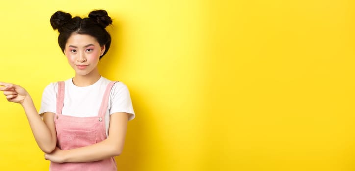 Beautiful teenage asian girl with bright makeup, pointing finger left at banner and smiling, yellow background.