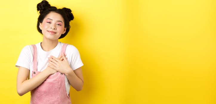 Beautiful teen asian girl in love, holding hands on heart and smiling touched, looking with affection, standing on yellow background.