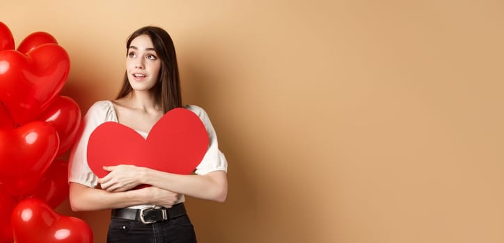 Romantic girl hugging Valentine day big red heart cutout, standing near romance balloons and looking left at logo, gazing dreamy at promo, beige background.