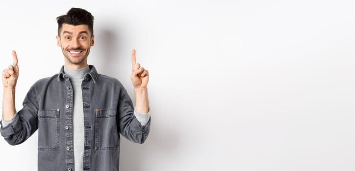 Excited young man smiling and showing advertisement, pointing fingers up at promo offer, standing on white background.