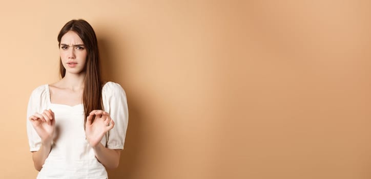 Stay away from me. Disgusted young woman frowning and looking with aversion at something stinky, rejecting disgusting thing, standing on beige background.