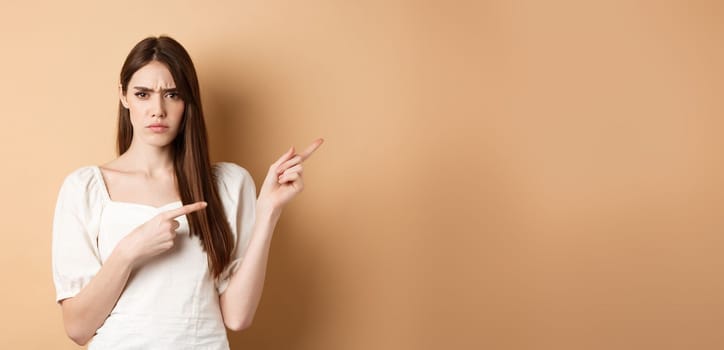 Angry young woman frowning, pointing fingers right and complaining, disappointed with logo, standing upset on beige background.