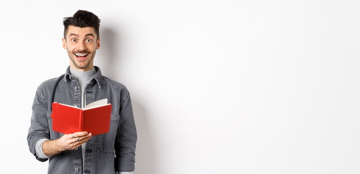 Hobbies and leisure. Happy young man reading planner, holding diary or red journal and smiling, making notes, standing on white background.
