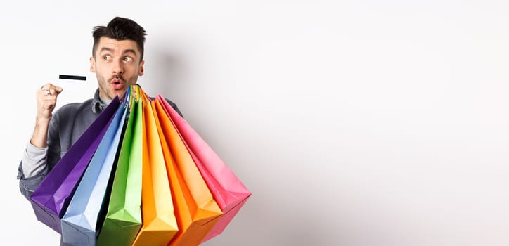 Excited young man buying in shop with plastic credit card, holding colorful shopping bags and looking aside at logo, standing against white background.