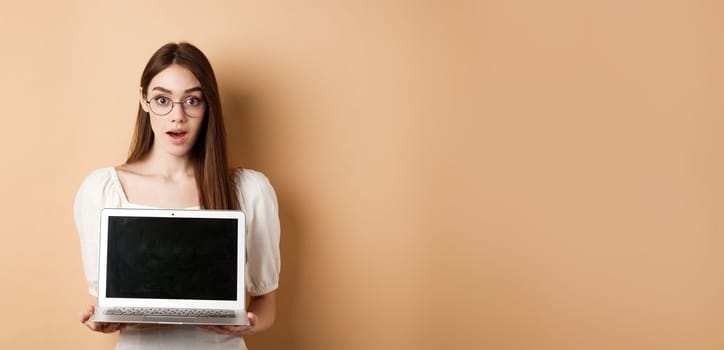 Surprised young woman in glasses showing laptop screen with amazed face, standing on beige background.