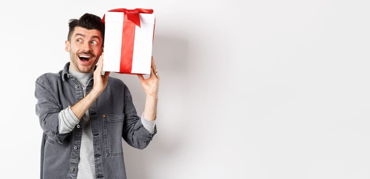Happy boyfriend shaking his gift to guess what inside, receive surprise present on Valentines day, smiling and looking aside cheerful, standing on white background.