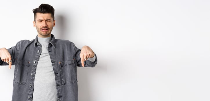 Sad crying man whining, pointing fingers down disappointed, showing bad news, standing upset on white background.