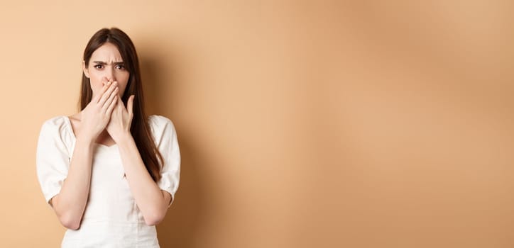 Shocked and confused woman frowning upset, covering mouth with hands, disappointed from something terrible, standing on beige background.