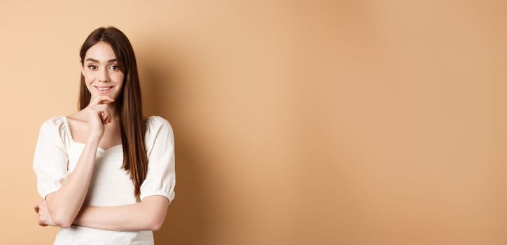 Smiling young european woman in dress, having an idea, listening with interest, touching chin and looking at camera intrigued, standing against beige background.