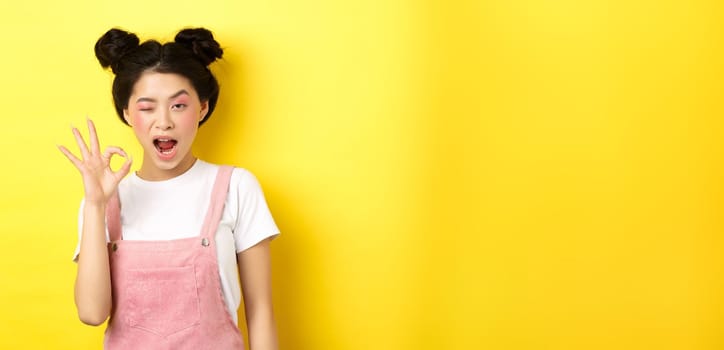 Alright. Confident asian female model with bright makeup and summer clothes, showing okay sign, winking and say yes, agree and approve, yellow background.