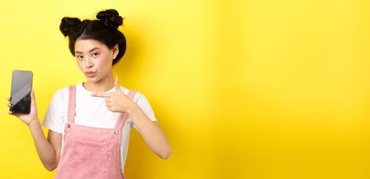 Online shopping concept. Stylish asian woman with pink makeup, pointing at empty smartphone sreen, showing good deal, yellow background.