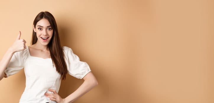 Amazed young woman showing thumb up, say yes and praise great product, approve choice, standing satisfied on beige background.