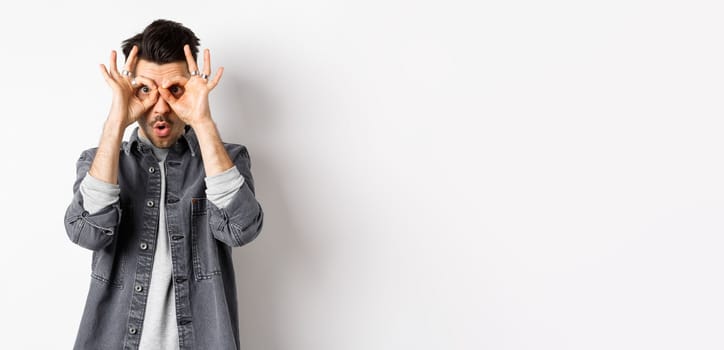 Funny guy look through hand binoculars, checking out awesome promo, say wow and stare at camera excited, standing on white background.