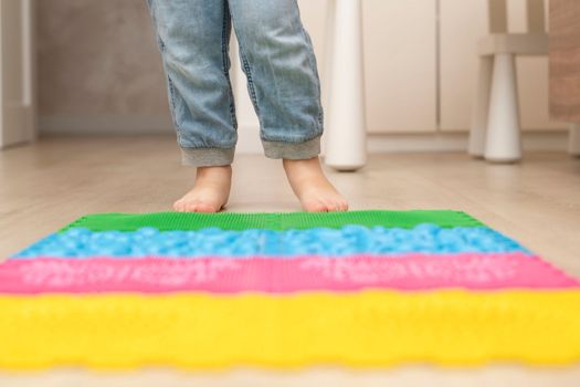 Health concept. A little boy stands with bare feet on a multi-colored orthopedic massage mat in a home interior. Close-up of children's heels. Back view.