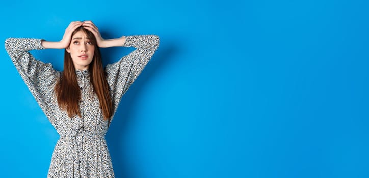 Troubled young woman with long hair, holding hands on head in panic, looking up nervous, having problem, standing on blue background.