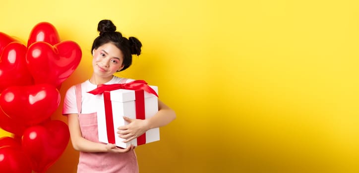 Happy Valentines day. Cute asian girl hugging surprise gift from boyfriend, standing near lovely red heart balloons and yellow background.