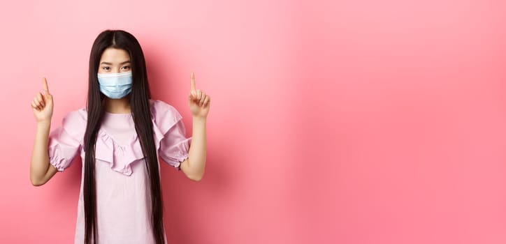 Coronavirus, quarantine and lifestyle concept. Beautiful asian woman in sterile medical mask pointing fingers up, smiling with eyes and look confident, standing against pink background.