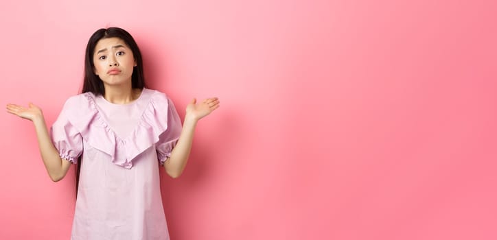 Sorry cant help. Clueless asian girl shrugging with sad face, know nothing, standing indecisive against pink background.