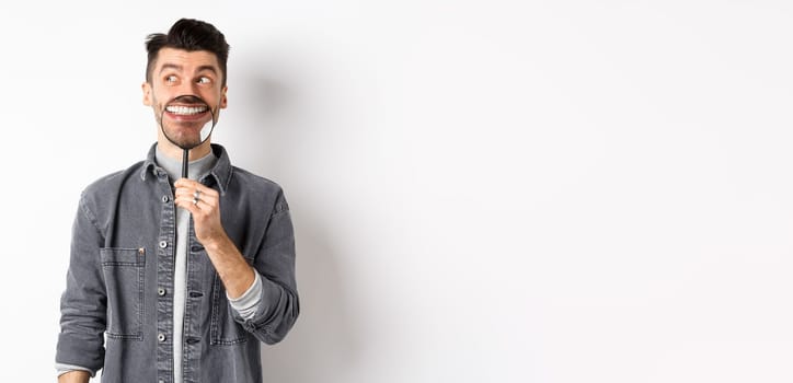 Handsome positive guy showing white perfect smile with magnifying glass, looking left at logo, standing against white background.