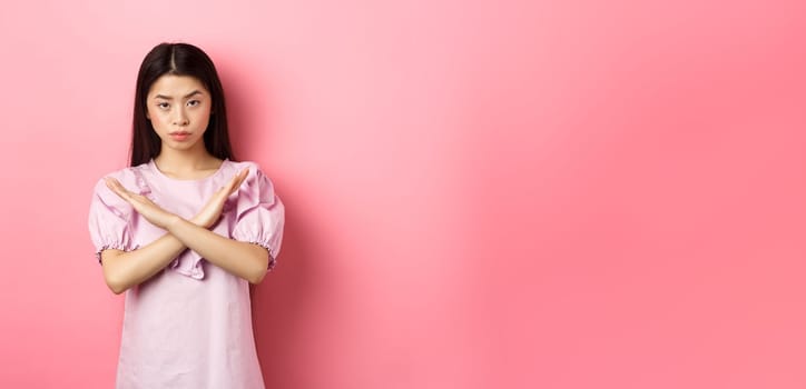 Serious chinese girl show cross sign, make stop gesture and look confident, block person, say no and reject offer, standing on pink background.