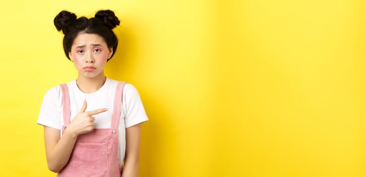 Sad and disappointed asian girl frowning, feel unfair, pointing finger right at bad thing, complaining on something upsetting, yellow background.