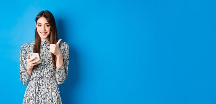 Happy smiling girl in stylish dress, showing thumb up in approval, using smartphone app and recommend it, standing against blue background.