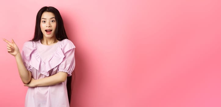 Excited beautiful asian teen girl pointing finger left, open mouth amazed and looking at camera, showing cool promo deal, pink background.