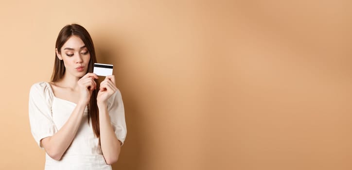 Cute young woman looking thoughtful at plastic credit card, thinking of shopping, standing on beige background.