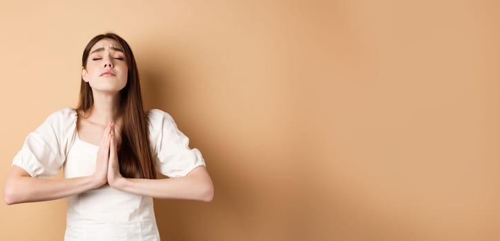 Hopeful woman close eyes and pray god, holding hands in begging gesture and making wish, say please, standing on beige background.