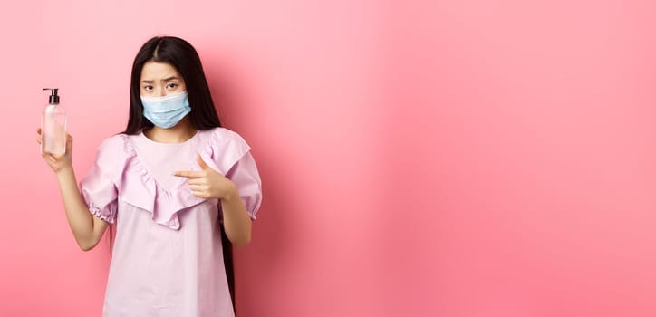 Healthy people and covid-19 pandemic concept. Skeptical asian woman frowning, wearing face mask, pointing at bottle of hand sanitizer, standing against pink background.