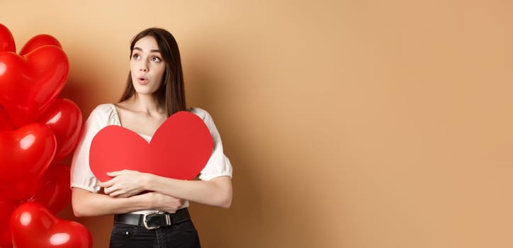 Valentines day and love concept. Intrigued tender girl hugging big red heart cutout and stand near balloons, looking aside at logo, saying wow, beige background.