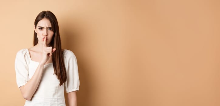 Angry young girl scolding bad behavious, shushing and frowning mad, tell to be quiet, looking displeased, standing on beige background.