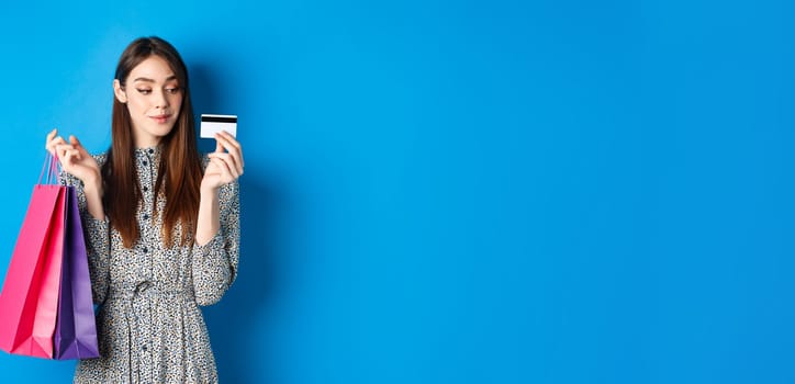 Beautiful girl going on shopping with plastic credit card, holding paper bags with purchased items, standing pleased on blue background.