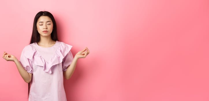 Stay calm. Peaceful asian woman meditating, hold hans in zen gseture and close eyes, practice yoga, standing patient on pink background.