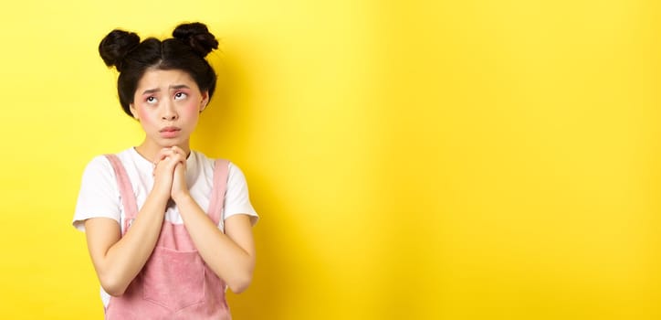 Sas asian girl holding hands in pray, pleading god, looking up and begging, standing on yellow background.