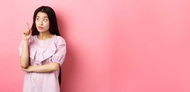 Beautiful asian girl raising finger and looking left at logo, having an idea, found solution, standing against pink background.