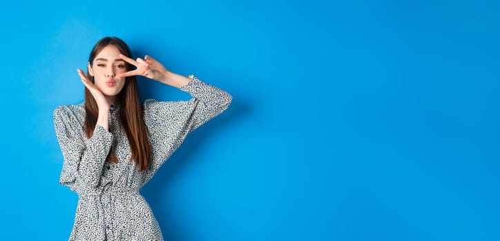 Coquettish young woman in dress showing v-sign on eye and pucker lips for kiss, standing happy on blue background.