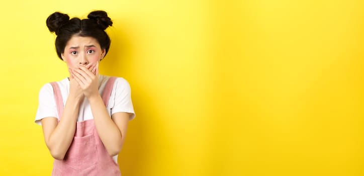 Worried asian teen girl covering mouth with hands, looking concerned and anxious, standing scared on yellow background with glam makeup and summer clothes.