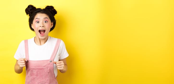 Excited asian woman open mouth, looking fascinated and amazed, receive good news, standing in summer clothes on yellow background.